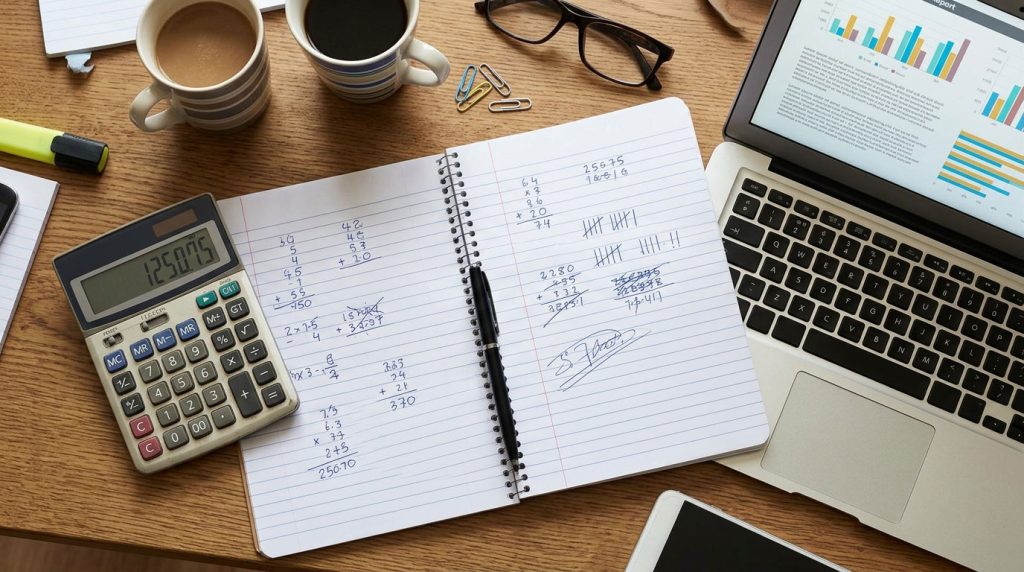 Notebook with handwritten calculations beside a news report on a laptop.